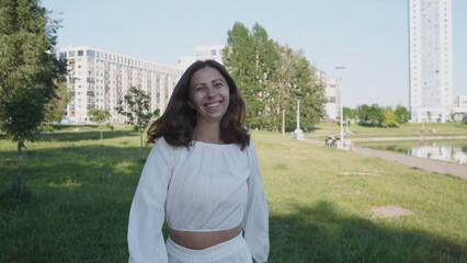 Young woman with long hair enjoys a sunny day in a park, wearing a white outfit and beaming with happiness as she strolls through the greenery.