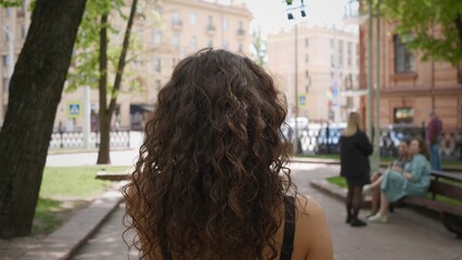 A young woman with curly hair walks through a bustling city park, enjoying the lively surroundings as friends gather nearby on a sunny afternoon in the city center.
