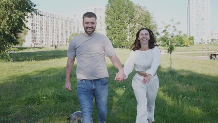 A joyful couple strolls through a vibrant park in the city, holding hands and enjoying the sunshine, while a small dog accompanies them along the path.