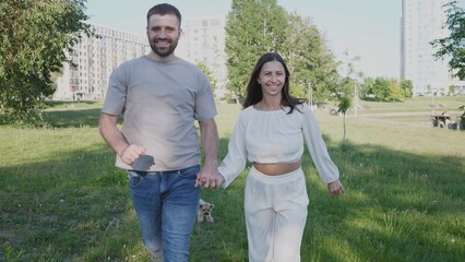 A joyful couple walks hand in hand through a grassy park, smiling and enjoying their time together with a small dog beside them on a sunny day.