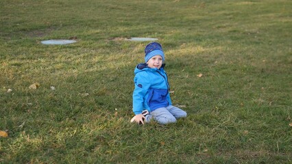 A smiling child sits on the grass, dressed in a blue jacket and hat, enjoying outdoor play in a park during a sunny autumn afternoon.