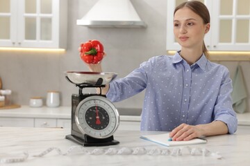 Woman weighting bell pepper on kitchen scale at white marble table indoors