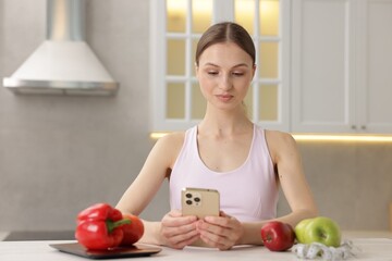 Woman using smartphone at white table with kitchen scale, healthy food and measuring tape indoors