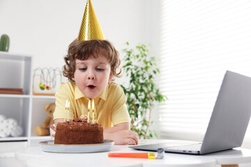 Birthday celebration. Little boy blowing candles on cake while having video call via laptop at...