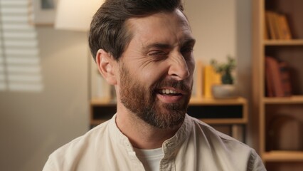 Fototapeta premium Cheerful Caucasian man joyful happy adult bearded male portrait at home weekend tenant homeowner blogger businessman guy looking at camera flirt wink one eye friendly emotion happiness toothy smile