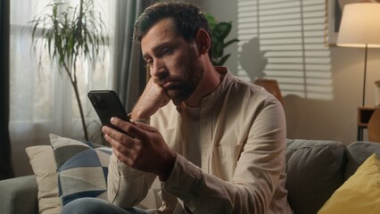 Fototapeta premium Sad upset frustrated man Caucasian businessman guy bored using mobile phone on couch sofa worried disappointed male homeowner reading news cellphone browsing message scam scrolling smartphone at home