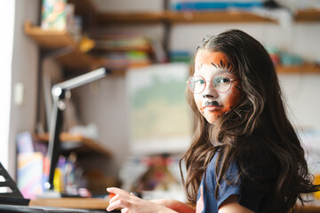 10-year-old girl with animal face paint and glasses sitting at piano, looking at camera. Concept:...