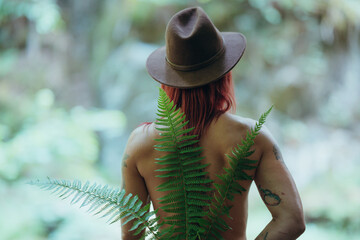 Serene redhaired woman with hat and green fern leaves posing in natural setting from behind