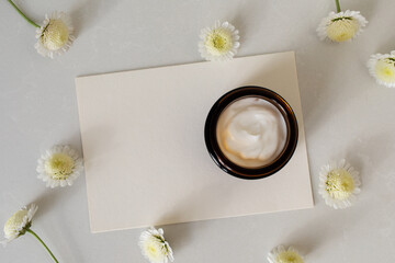 Jar of cosmetic cream and blank paper card on stone table decorated with flowers. Beauty, spa and skincare lifestyle aesthetic concept top view.