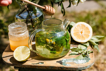 Close-up of female hands adding honey to a clear teapot with herbal infusion in garden light. Warm and calming ritual.