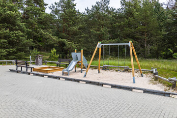 Fototapeta premium Empty Forest Playground with Wooden Swing Set, Metal Slide and Sandbox on Paved Park Surface