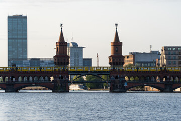 Fototapeta premium Wide view of Oberbaumbrucke in Berlin with a yellow U-Bahn train crossing above the river. The twin brick towers and modern skyline highlight the city s architectural contrast.