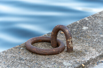 Rusty iron mooring ring fixed in a concrete quay beside calm blue water, creating a serene maritime...