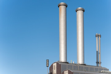 Two large industrial chimneys rise into the blue sky. The modern facility dominates the cityscape with its vertical architecture.