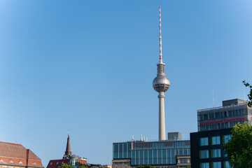 The TV Tower dominates the cityscape alongside modern office buildings and a historic church steeple. Clean lines and a blue sky characterize this urban scene.