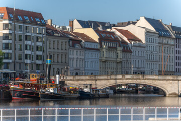Historic buildings and houseboats line the riverbank in front of an old stone bridge in Berlin. An idyllic scene of urban waterways in sunlight.