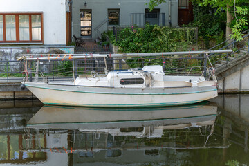 A white sailboat rests peacefully in the water in front of a residential building. The reflection in the water lends the image a tranquil atmosphere.