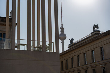 The Berlin TV tower is visible through modern columns, next to it are historic museum buildings with roof sculptures. The perspective emphasizes the contrast between old and new.