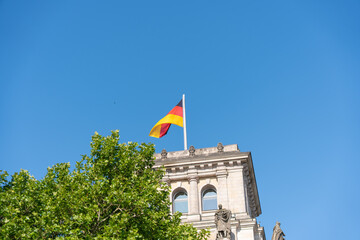 The German national flag waves atop a historic building with statues and columns. Bright blue sky and green tree leaves frame the scene.