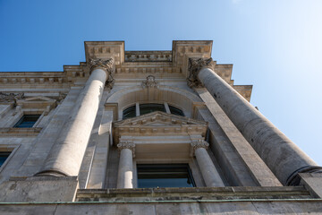 Neoclassical Facade with Columns. Upward view of a neoclassical building with large stone columns and ornate details. The clear blue sky frames the monumental structure.