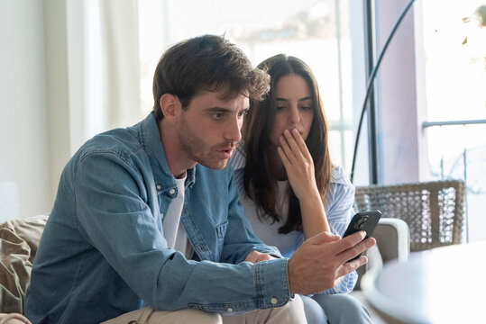Worried young couple at home looking at smartphone with serious expressions. Facing bad news, possible online scam, job-related issue or financial concern. Real-life anxiety, uncertainty and digital s