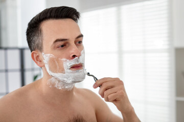 Handsome man shaving with razor near mirror at home