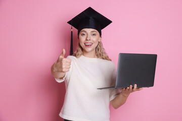 Happy student with laptop showing thumbs up after graduation on pink background