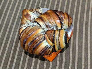 Four freshly baked marbled chocolate buns are arranged in an orange and white paper box on a brown and cream striped cloth surface, showing off the soft texture and tempting chocolate coating.