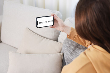 Cyber bullying. Upset teenage girl looking at smartphone with text You Loser on sofa at home,...
