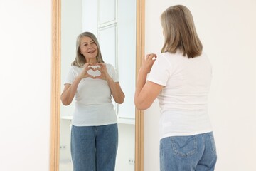 Beautiful senior woman making heart gesture near mirror indoors