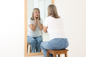 Beautiful senior woman looking at herself in mirror indoors