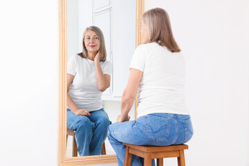 Beautiful senior woman looking at herself in mirror indoors