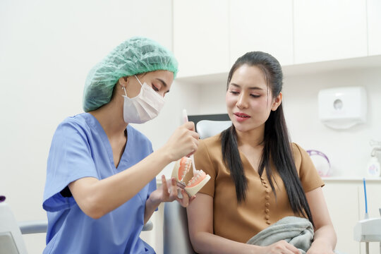 asian female dental assistant in scrub mask demonstrating proper tooth brushing technique using toothbrush and dental model to asian female patient during consultation in dental clinic
