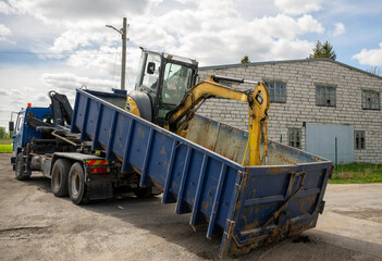 Blue industrial truck carrying a yellow mini excavator in its container bed 