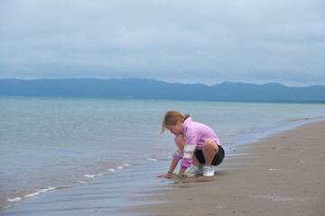 Young child crouched on a serene beach, intently playing with sand against the backdrop of gentle waves and distant mountains under a cloudy sky,