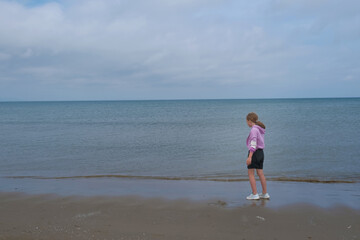 Young girl in a pink hoodie and shorts stands quietly at the water's edge, gazing at the vast sea under a cloudy sky.