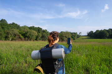 An Asian woman uses a smartphone navigation app while hiking with a backpack equipped with a solar panel charger, reflecting sustainable trekking, off-grid adventure, and eco-friendly travel.