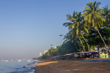 Tropical morning at Jomtien Beach with calm sea, leaning coconut palms over sun umbrellas on golden...