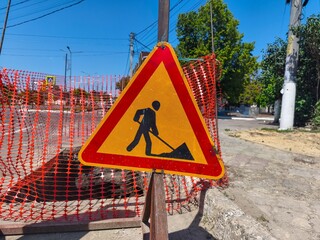 A construction worker is digging a hole in the street. A yellow and red sign warns drivers to slow...
