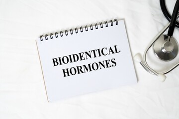 The medical concept "BIOIDENTICAL HORMONES" is written clearly on a white spiral notebook, accompanied by a stethoscope on a white background.