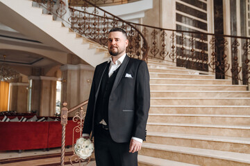 A groom in a black suit with a bouquet is waiting for the bride at the stairs