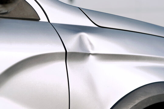 Deep dent on the front fender of silver car, closeup. Car surface damage, impact marks, and deformation of the vehicle body panel. Silver car with visible damage and pronounced dent on the fender