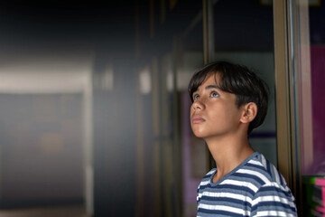 A contemplative teenage boy in a striped blue shirt looks upward indoors, standing near a glass...