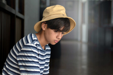A contemplative teenage boy in a striped blue shirt looks down indoors, sitting near a glass wall with soft natural light. Mood of hope, thought, detention, lonely, sad, broken family and solitude.