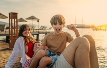 Young happy Caucasian family sitting with son boy on sea pier at sunset, vacation summer travel concept