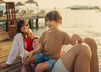 Young happy Caucasian family sitting with son boy on sea pier at sunset, vacation summer travel concept