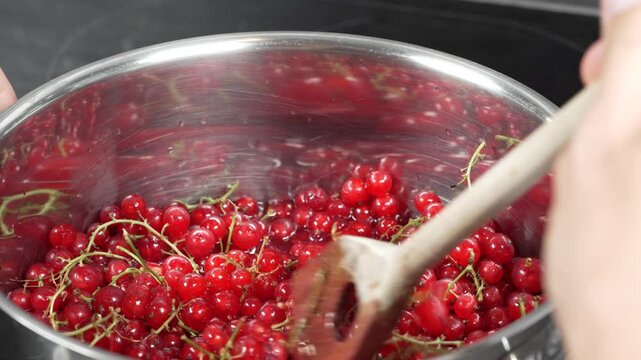 Red currants in a pot are crushed with a wooden spoon and release juice to make jelly