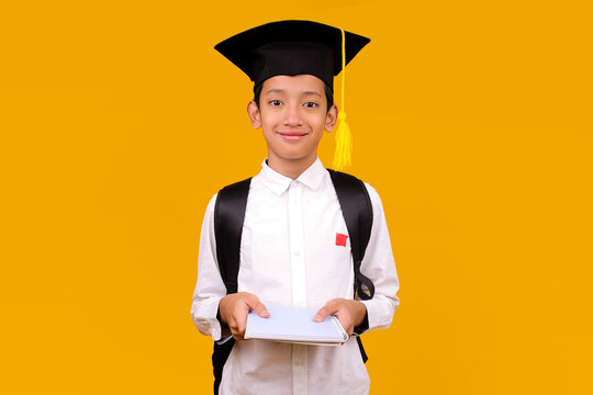 Positive Confidently Young Student Boy Wear Graduation Cap Holding Notebook On Yellow Background - Powered by Adobe
