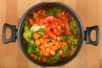 A top-down view of a cooking pot with chopped tomatoes, carrots, and green leafy vegetables in water, ready for cooking
