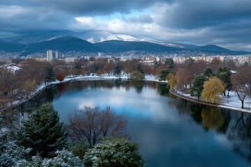 Fototapeta premium Lake reflects sky snowy mountains behind city buildings winter scene with trees snow patches